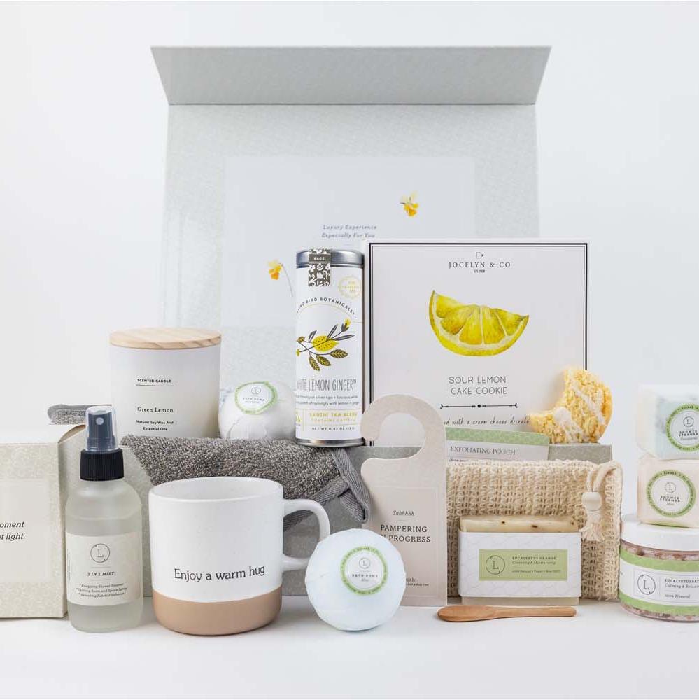Get-well gift basket designed to promote mindfulness and wellness, helping to bring calm and comfort during recovery. The set includes a ceramic cup, a scented candle, eucalyptus and mint shower steamers, mint and eucalyptus bath bombs, eucalyptus soap, a large jar of eucalyptus salt, tea bags, lemon cookies, a soap saver, mist, a gray towel, and a door hang tag. All items are elegantly arranged in a beautiful gift box that can be personalized with the recipient's name in rose gold lettering for a special