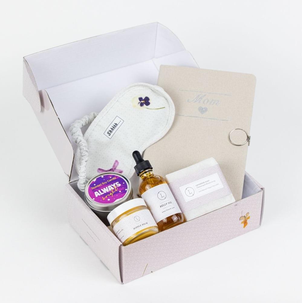 Pregnancy care package thoughtfully designed for a new mom, featuring essential items that are both practical and pampering. The set includes nipple balm, belly oil, a scented soy candle in a tin, a 'Mom' journal with a heart symbol, lavender natural soap, and a cotton eye mask. All items are beautifully arranged in a customizable gift box, with the option to add the recipient’s name on the lid for a personalized touch.