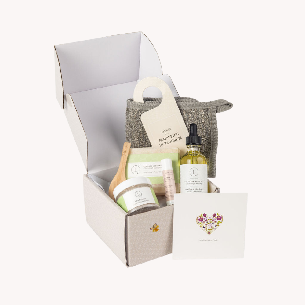 Natural skincare gift set by Lizush designed for relaxation and rejuvenation, featuring handmade products with 100% natural ingredients. The bath gift set includes lavender body oil, foot salts, a lemongrass natural soap bar, unscented lip balm, a charcoal washcloth, and a door hang tag with the message 'Pampering in service.