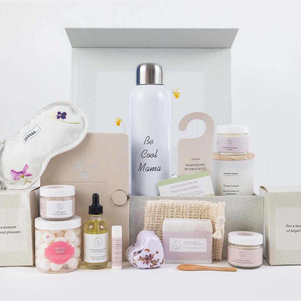 Mother's gift basket designed to celebrate and honor mothers, featuring a curated selection of pampering and self-care items. The set includes a journal with 'Mom' and a heart symbol, a water bottle, a scented candle, lavender shea butter, lavender body oil, lavender foot salt, a heart-shaped lavender shower steamer, lavender natural soap, a facial clay mask, candy sparkle, a cotton eye mask, a natural luffa soap saver, and an unscented lip balm. The basket also comes with a door hang tag. All items are bea