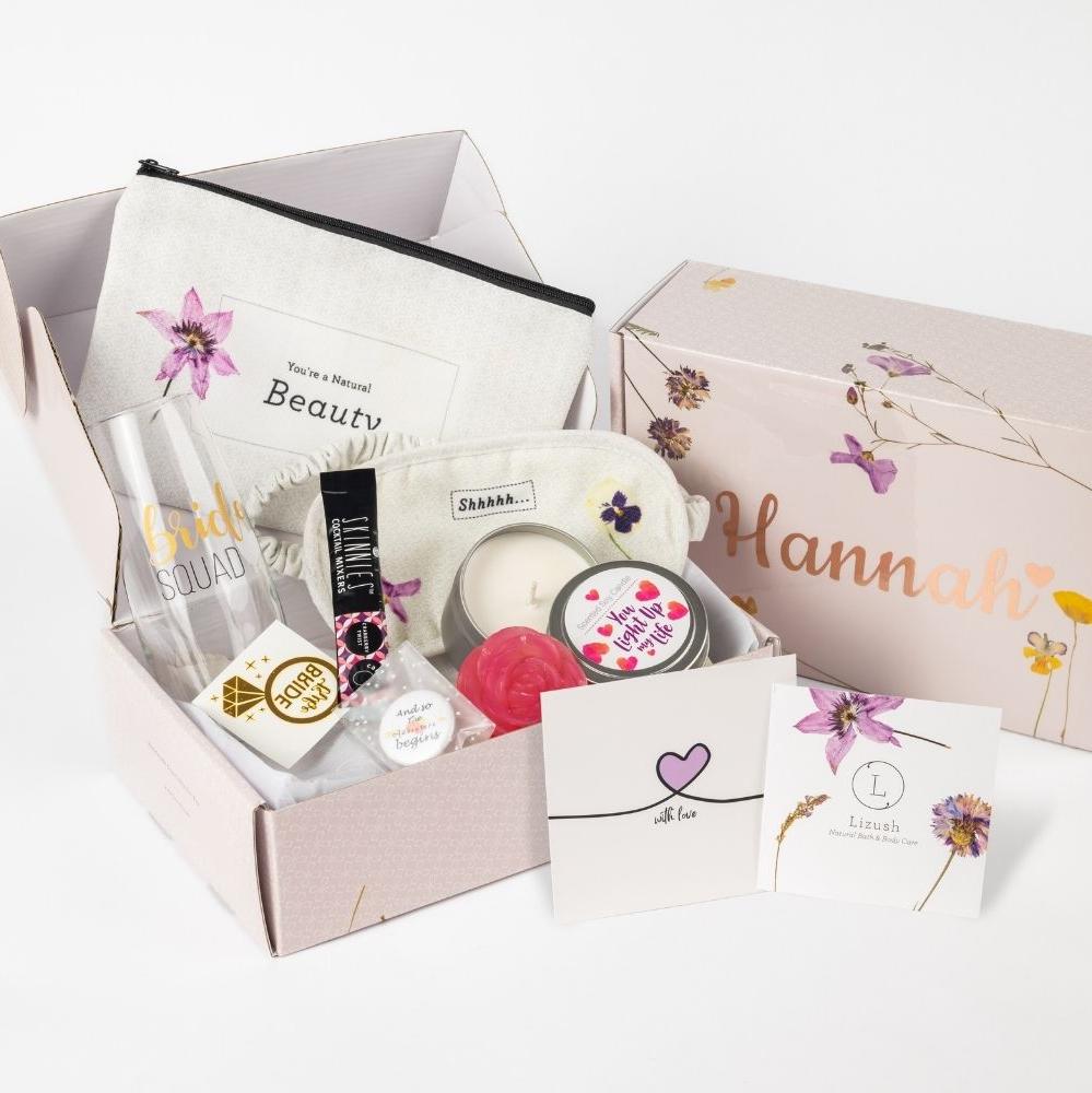 Bridesmaid Spa Gift Set displayed with luxurious self-care and pampering items, ideal for a bridal shower or bridesmaids. The set includes a long glass champagne cup, a soap flower, Skinnies drink, a pin with the phrase 'and so the adventure begins...', a temporary tattoo, a cosmetic bag, a candle adorned with hearts, a purple greeting card, and an eye mask. All items are elegantly arranged in a beautiful box, which can be personalized with the recipient’s name in rose gold lettering. A personal note can al