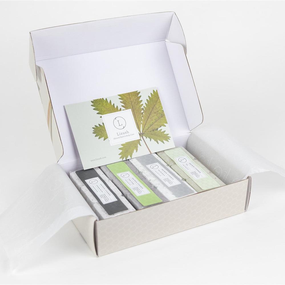 Image of a luxury shower steamer gift set displayed in an elegant rectangular box (8''x6''x3'') with a personalized name option on the lid. The set includes 12 handmade aromatic shower steamers arranged neatly, featuring four scents: Lemongrass Tea Tree, Cedar Orange, Pine, and Mint Eucalyptus. Each steamer has a rustic, organic look, with visible salt, petals, and seeds on top, highlighting the natural ingredients. 