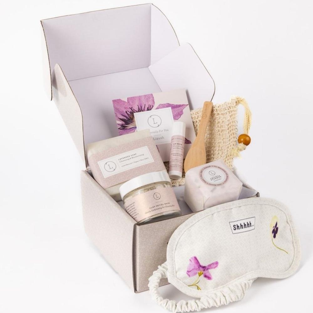 Small lavender gift box designed for expressing gratitude or showing appreciation, featuring a selection of relaxing skincare products. The set includes a 4oz lavender natural soap bar, a cotton eye mask, a 1oz clay facial mask, an unscented natural lip balm, a large shower steamer (2" diameter), and a natural luffa soap saver. All items are beautifully arranged in a customizable gift box, where the recipient's name can be added in rose gold lettering for a personal touch. A personalized greeting card is al