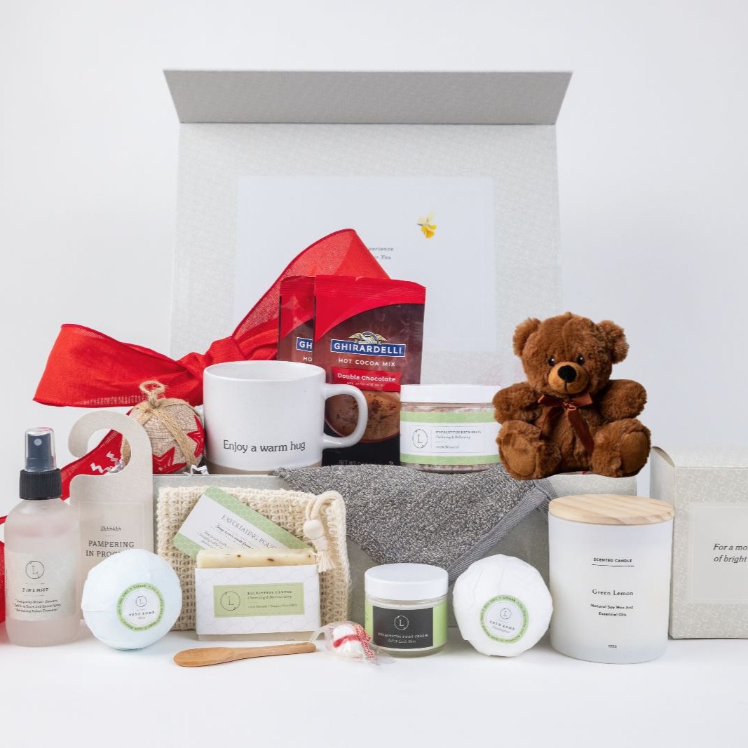 Valentine's Day gift basket designed to pamper and delight the special woman in your life, featuring a curated selection of luxurious and thoughtful items. The set includes a ceramic mug, a scented candle, eucalyptus bath salt, eucalyptus soap, eucalyptus and mint bath bombs, foot cream, mist, hot cocoa, a cute teddy bear, peppermint candies, a soap saver, and a door hang tag. All items are beautifully arranged in a gorgeous gift box that can be personalized with the recipient’s name in rose gold lettering 