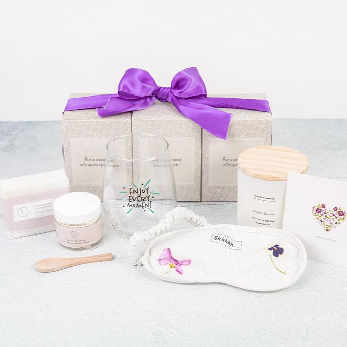 Lizush Spa Gift Box Set, an all-natural self-care package for women, perfect for relaxation and pampering. The set includes five pieces: an 'Enjoy every moment' wine glass, an 8 oz large candle with a wooden lid, a facial clay mask, natural lavender soap, and an eye mask. Packaged as a luxurious gift basket, it's ideal for mothers, daughters, wives, sisters, teachers, best friends, grandmothers, or for personal use. Made with 100% natural, eco-friendly, and recyclable materials, this aromatherapy bath gift 